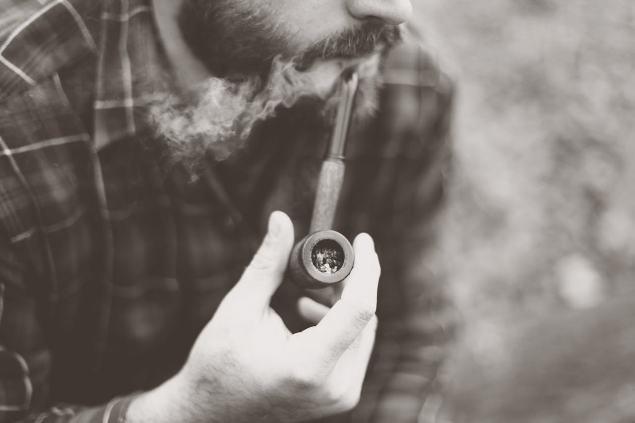 A close-up shot of a bearded man smoking a pipe outdoors.