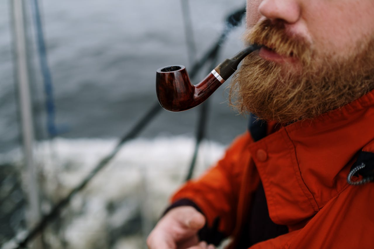 A bearded man in an orange jacket smokes a pipe on a sailboat during winter.