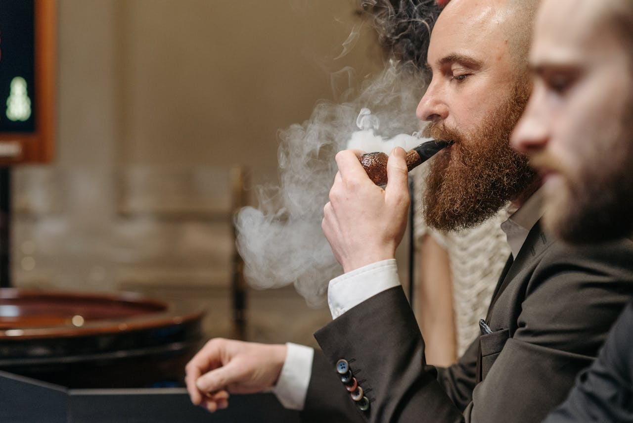 A bearded man in a suit enjoying a pipe indoors, surrounded by smoke.
