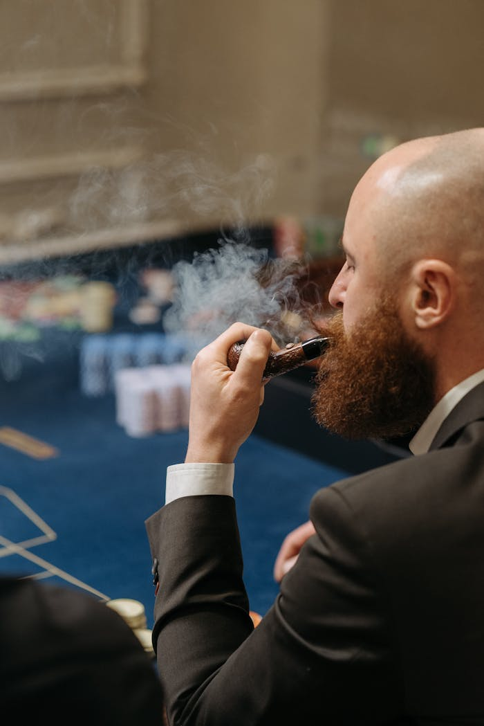 Bald man with beard smokes a pipe inside a casino, surrounded by a sophisticated atmosphere.