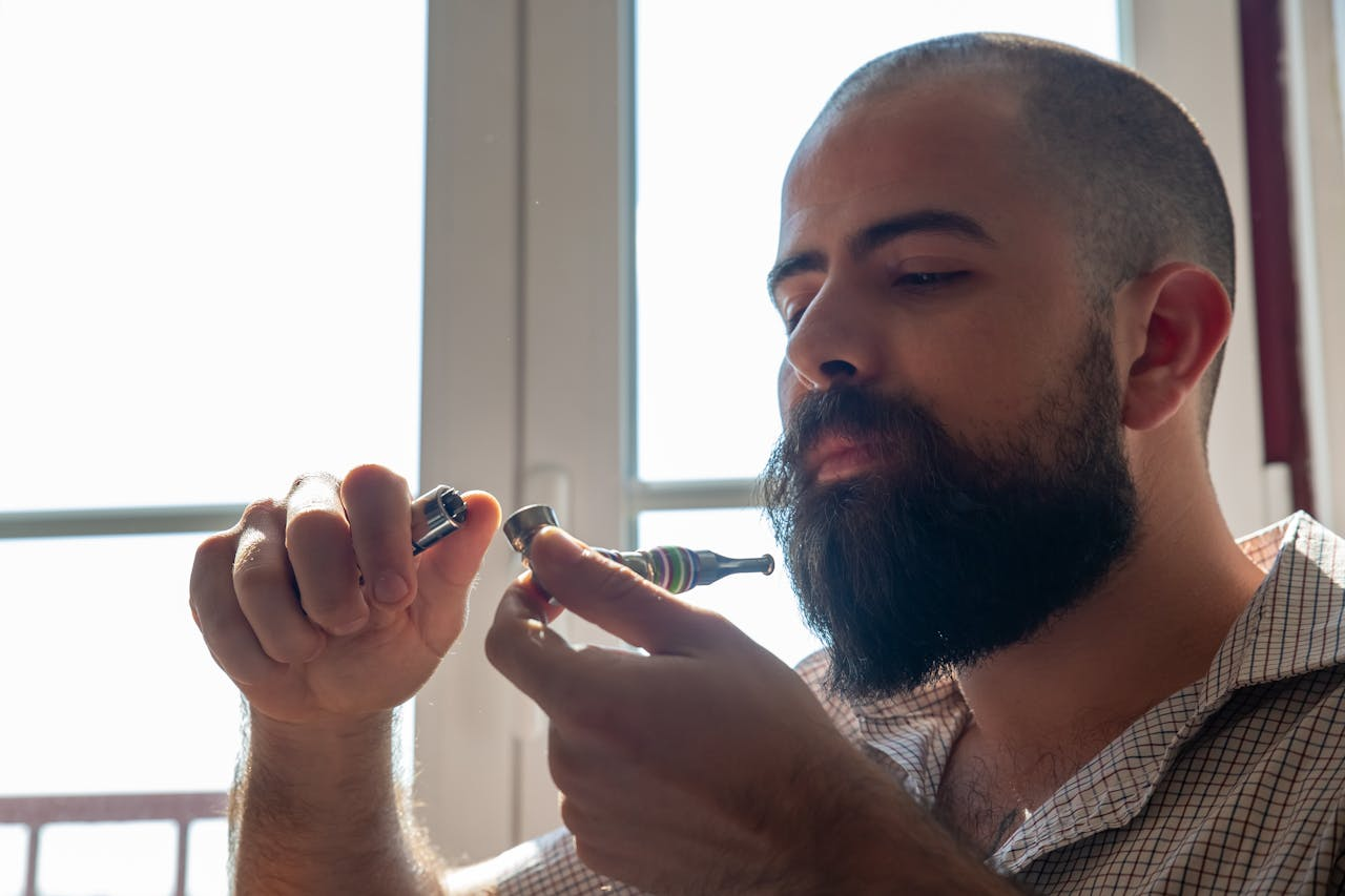 A bearded man in a plaid shirt lights a smoking pipe near a window, enjoying the moment.